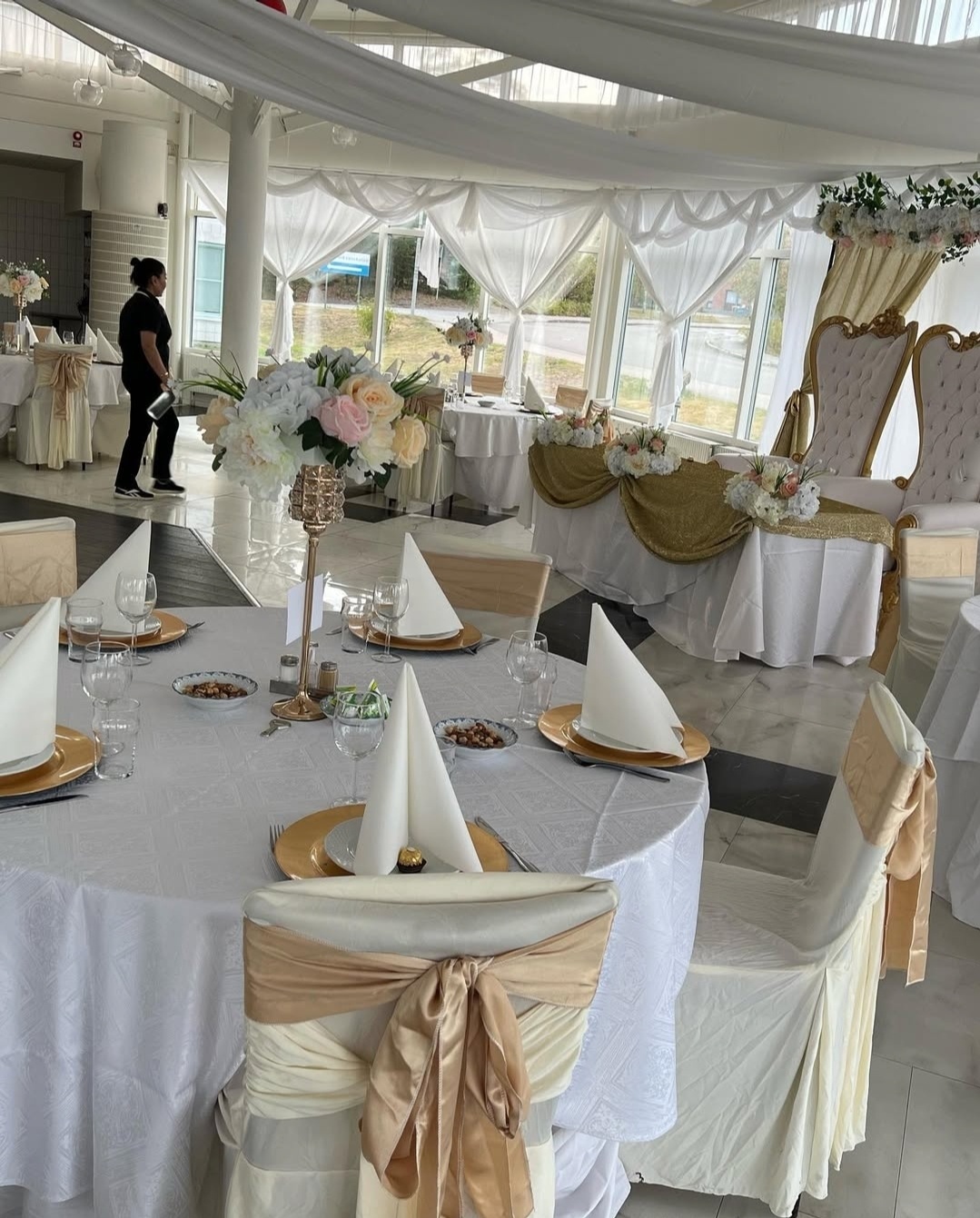 Champagnefärgade stolsrosetter, höga blomsterarrangemang och mjukt draperade tyger i taket skapar en lyxig och sammanhållen miljö.